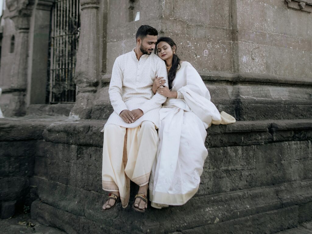 A couple in traditional white clothing sitting by a historic stone building outdoors.